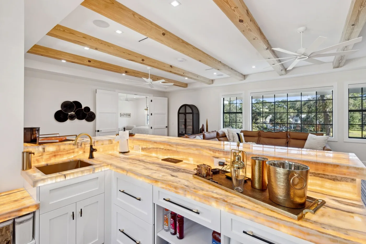 Home bar with a backlit onyx countertop, white cabinetry, and exposed ceiling beams by M&H Construction & Design