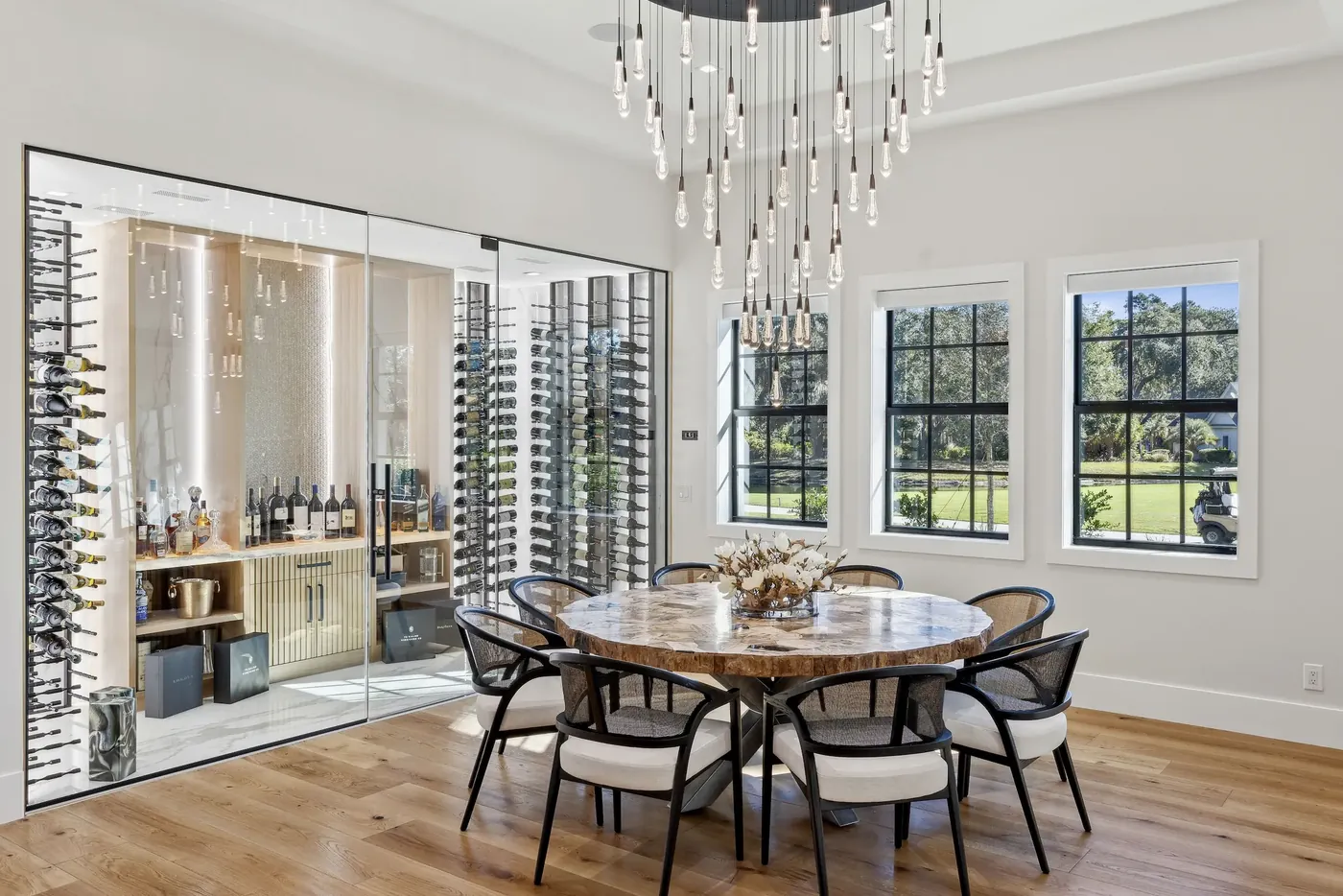 Dining room with a sculptural chandelier beside a glass-enclosed wine room in the M&H Timber & Tone project in Bluffton, SC